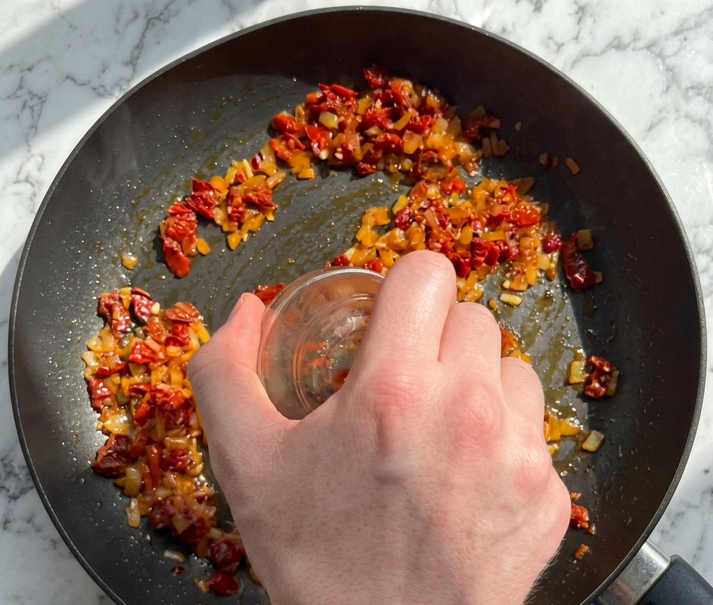 Une main versant des tomates séchées hachées et des flocons de piment rouge depuis un ramequin en verre dans la poêle daromates.
