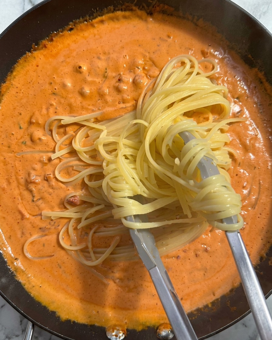 Un nid de pâtes cuites saisi à la pince et déposé dans une poêle remplie dune riche sauce crémeuse orangée.