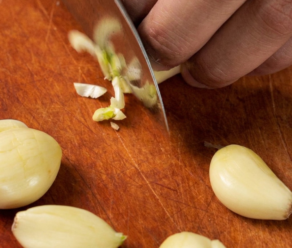 Gros plan sur un couteau de chef éminçant finement plusieurs gousses dail frais sur une planche à découper en bois.