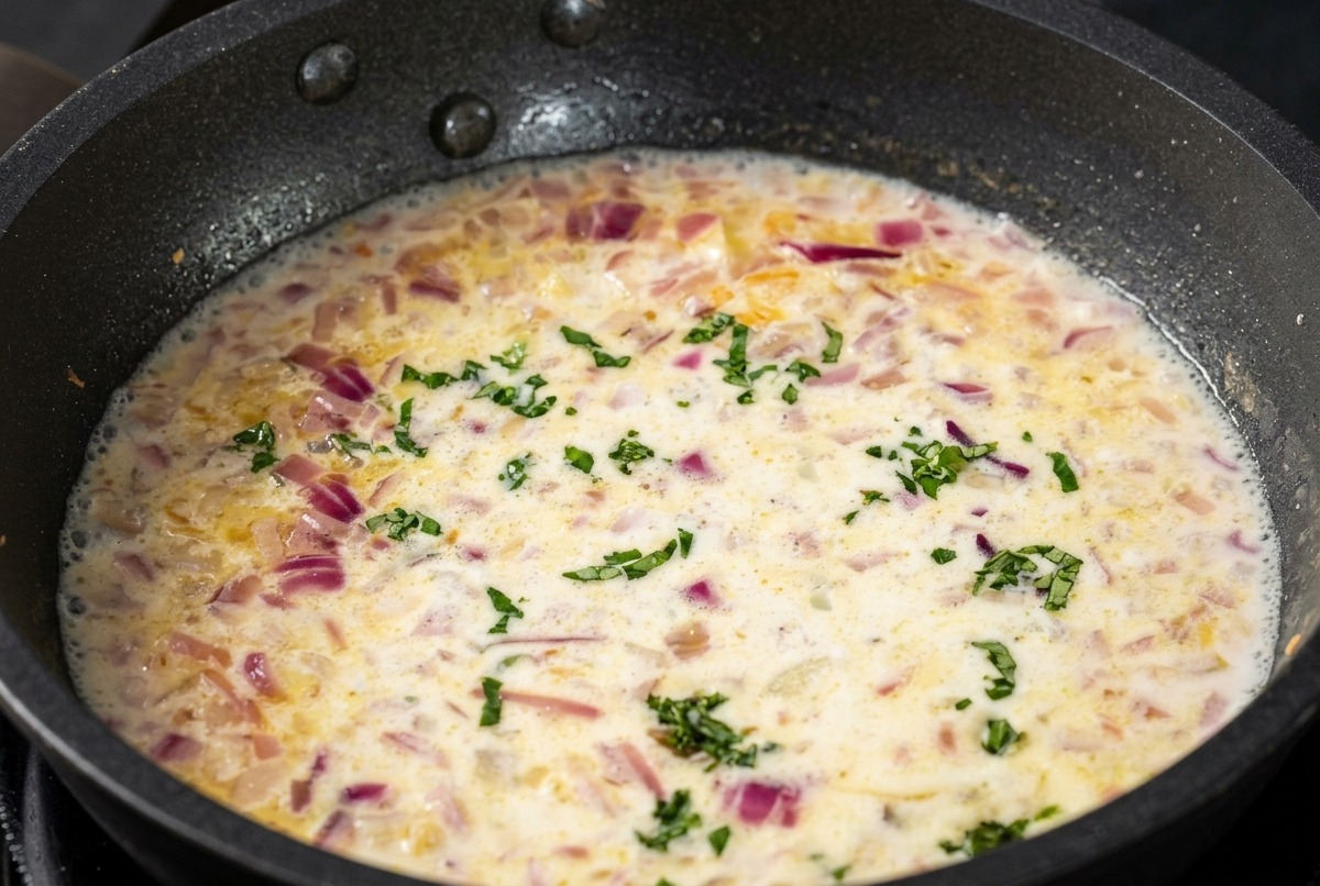 Une sauce crème blanche frémissant dans une poêle, parsemée déclats verts de basilic ciselé.