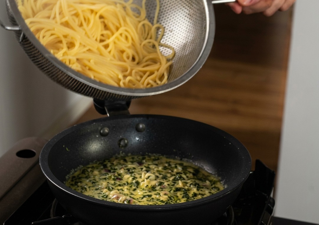 Spaghettis cuits versés depuis une passoire en inox dans une poêle de sauce crème parsemée de basilic.