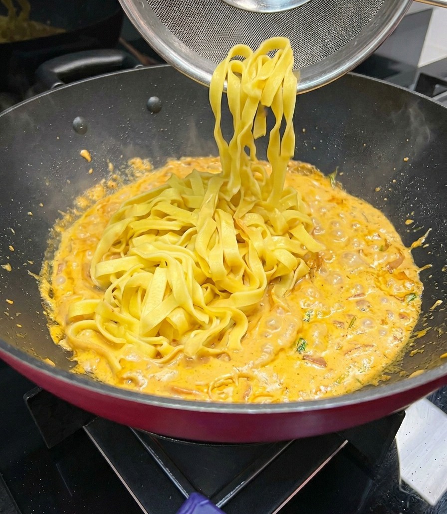 De larges bandes de fettuccine soulevées avec une passoire à mailles et ajoutées à une poêle de sauce crème orange qui mijote.