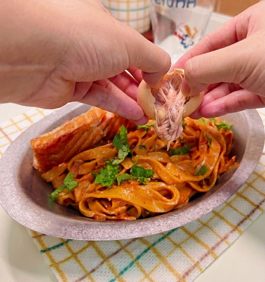 Des mains écalant délicatement et déposant un œuf mollet sur un plat de pâtes à la crème épicée et de saumon saisi.