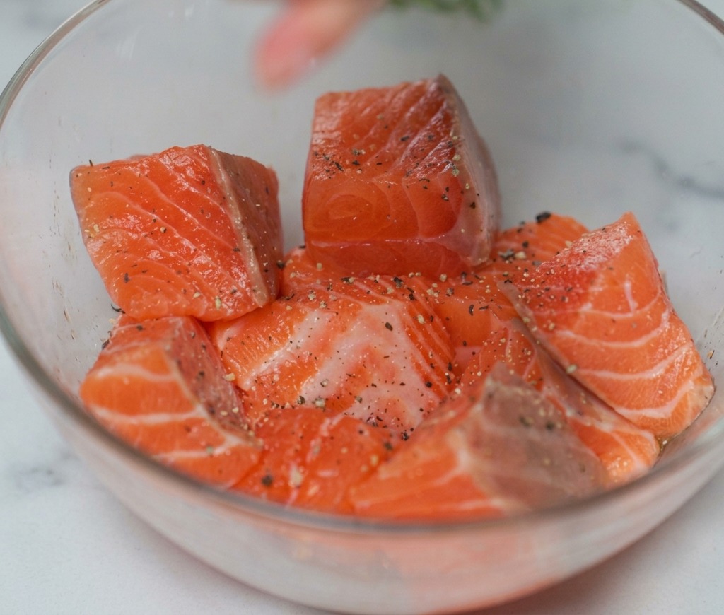 Dés de saumon cru dans un bol en verre, saupoudrés de sel et de poivre noir du moulin.