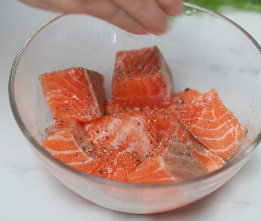 Une main pressant du jus de citron frais sur des dés de saumon assaisonnés dans un bol.