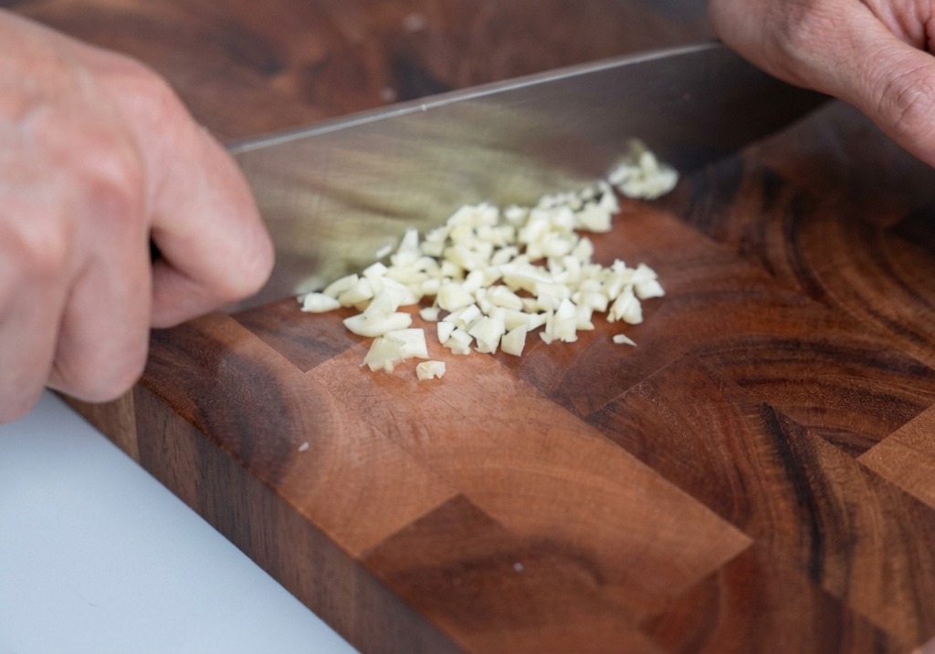Ail finement haché sur une planche en bois avec un couteau de chef posé juste à côté.