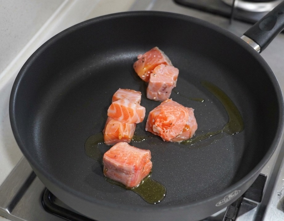 Dés de saumon en train de saisir dans une poêle antiadhésive noire avec de lhuile dolive sur un piano de cuisson à gaz.