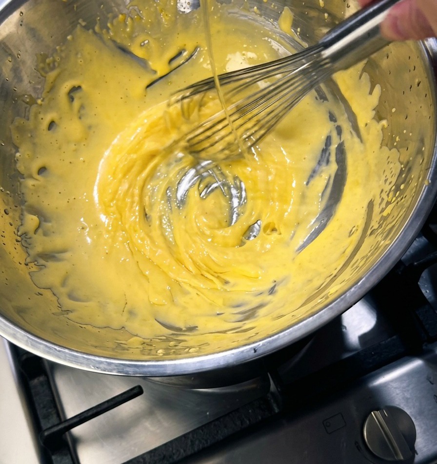 Un fouet en métal mélangeant une préparation épaisse jaune pâle dans un bol en inox, tandis quun fin filet de beurre doré y est versé.