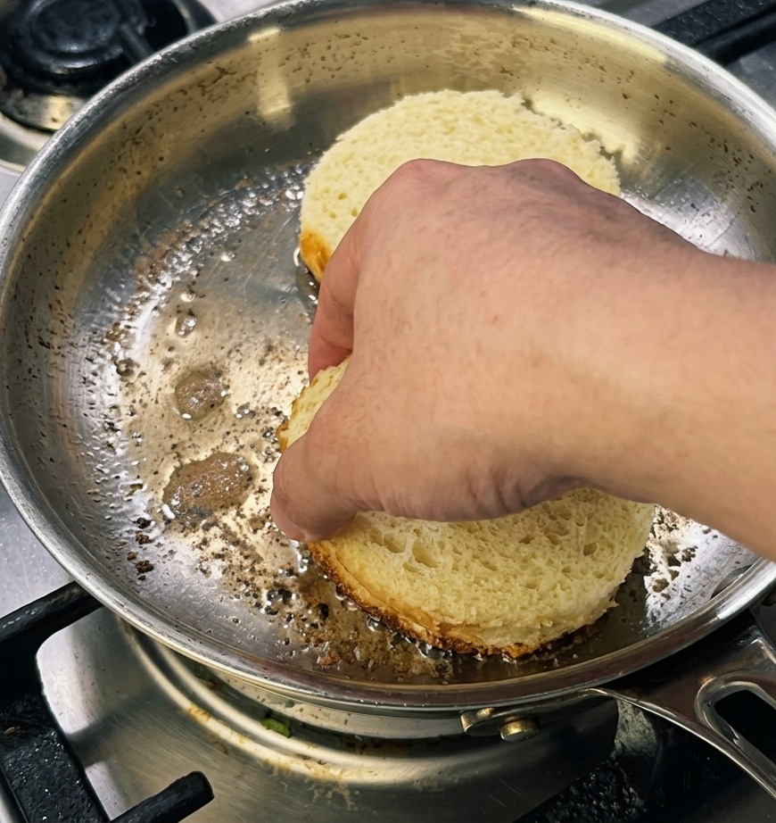 Une main pressant légèrement deux épaisses tranches rondes de pain brioché dans une poêle pour obtenir une croûte dorée.