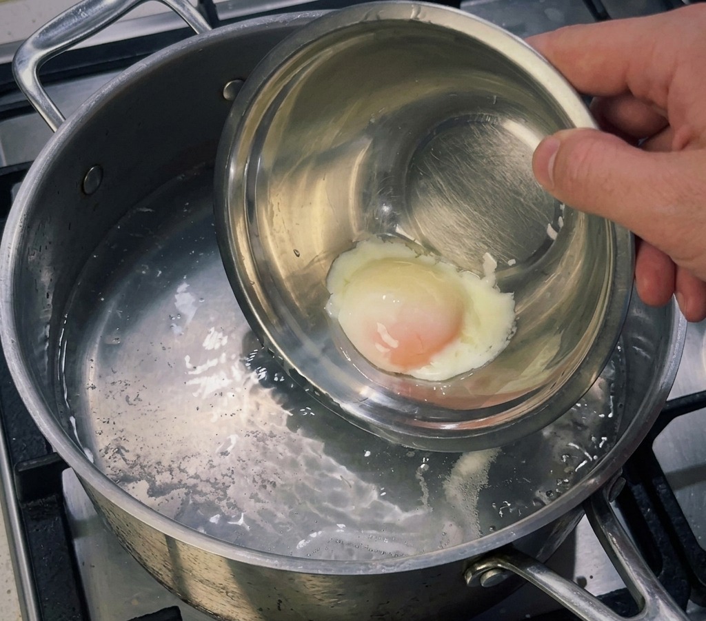 Un petit bol en métal contenant un œuf mollet délicatement plongé dans une casserole deau frémissante pour raffermir le blanc.