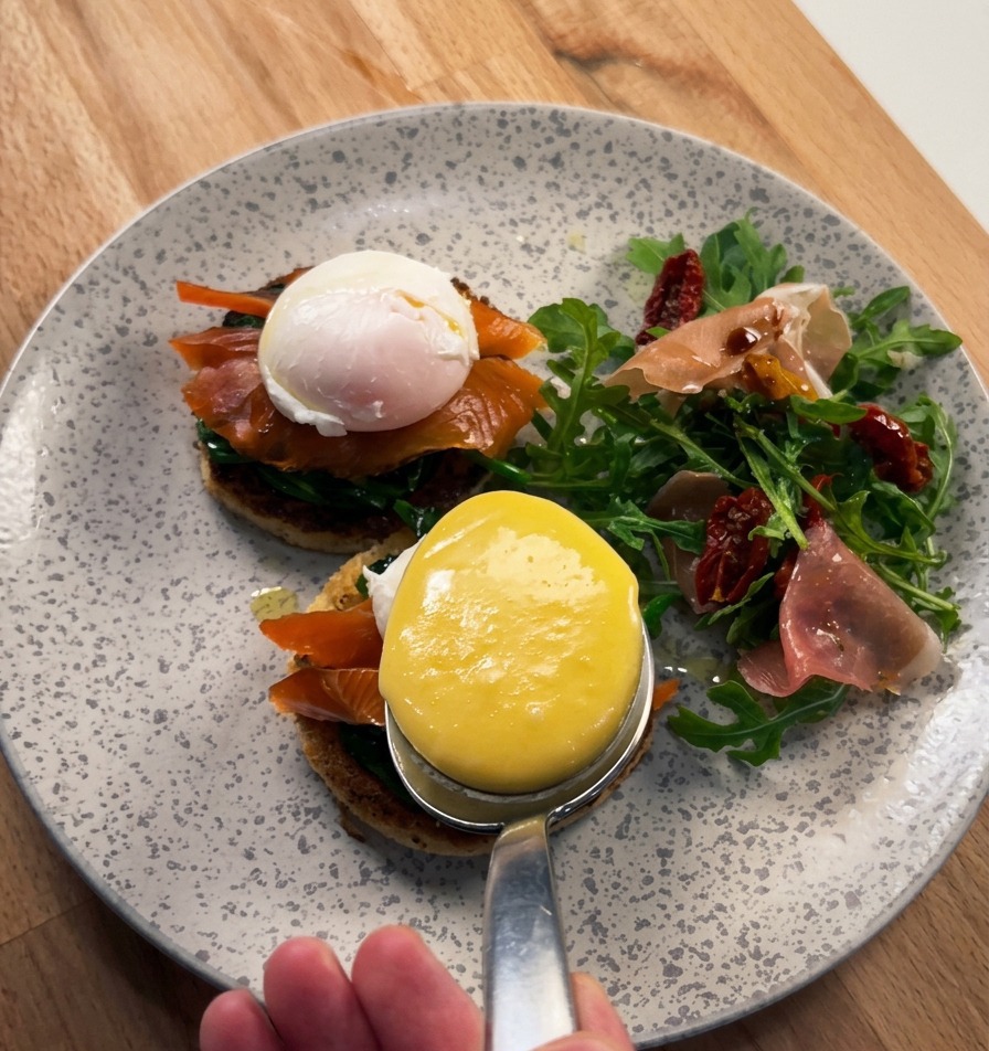 Gros plan sur une grande cuillère nappant une sauce hollandaise jaune et brillante sur un œuf poché posé sur du saumon fumé et une brioche.