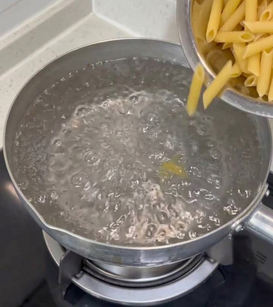 Des penne sèches versées depuis un bol en inox dans une casserole deau bouillante sur une plaque de cuisson.