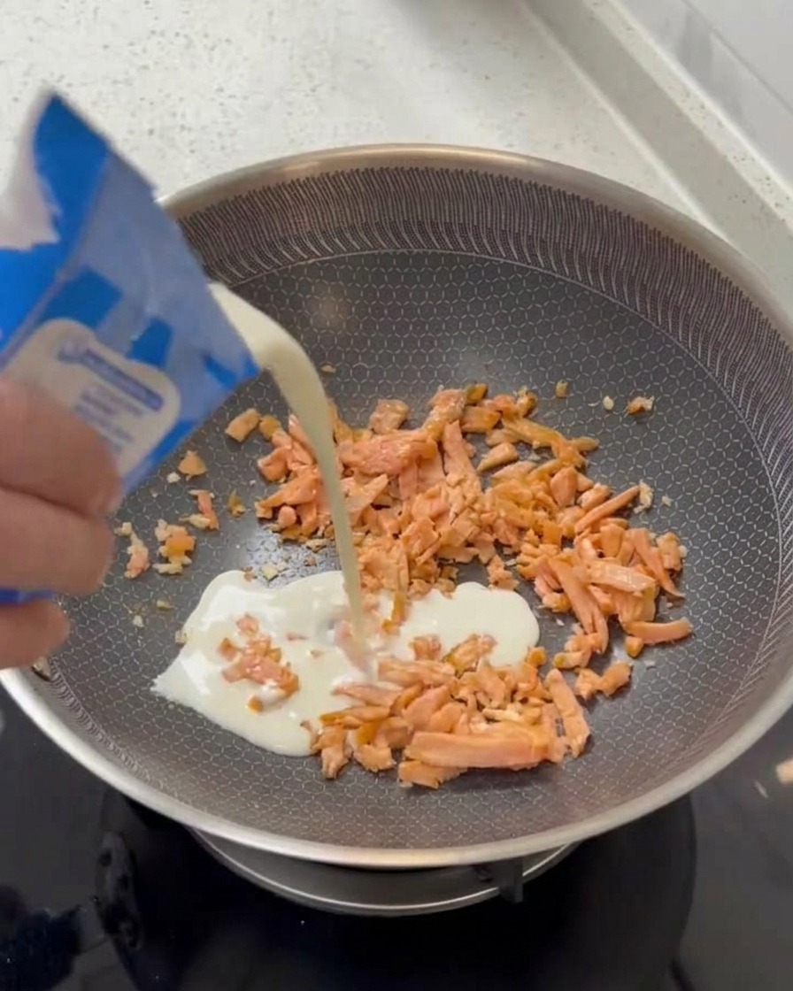 Une main versant de la crème liquide depuis une brique bleue et blanche dans une poêle avec le saumon.