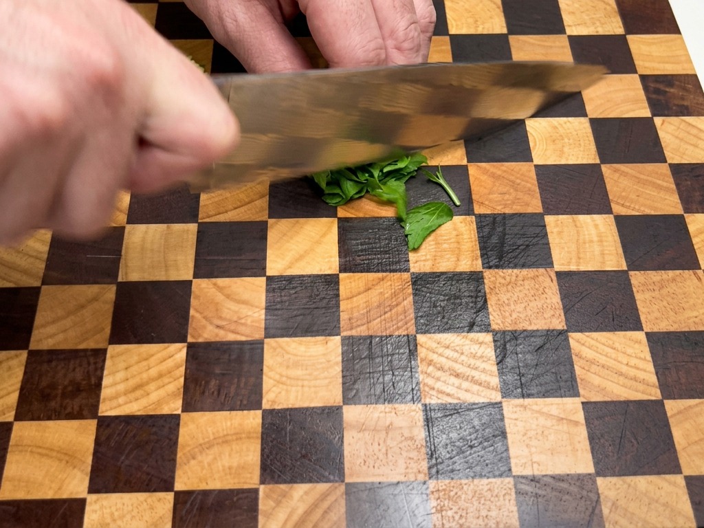 Gros plan sur des mains utilisant un couteau de chef pour hacher finement des feuilles de persil frais sur une planche en bois.
