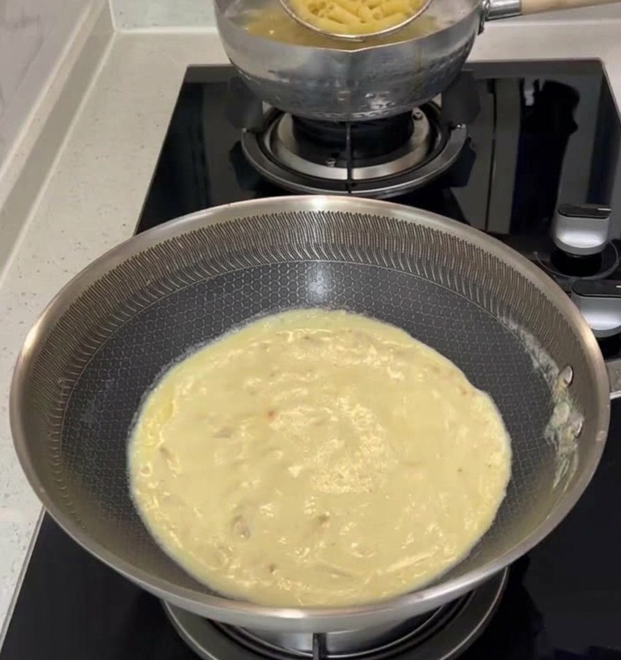 Une araignée de cuisine soulevant les penne cuites de la casserole deau bouillante pour les transférer dans la poêle de sauce crème.