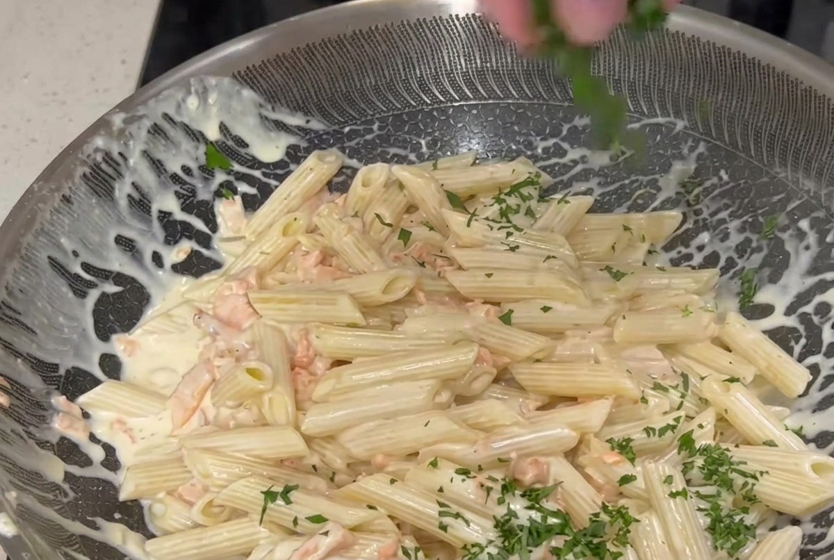 Une main parsemant du persil haché sur une grande poêle de penne à la crème et au saumon fumé.