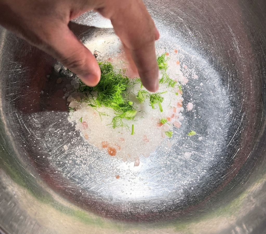 Une main mélangeant de laneth frais et des épices entières à du sel et du sucre dans un bol en inox.