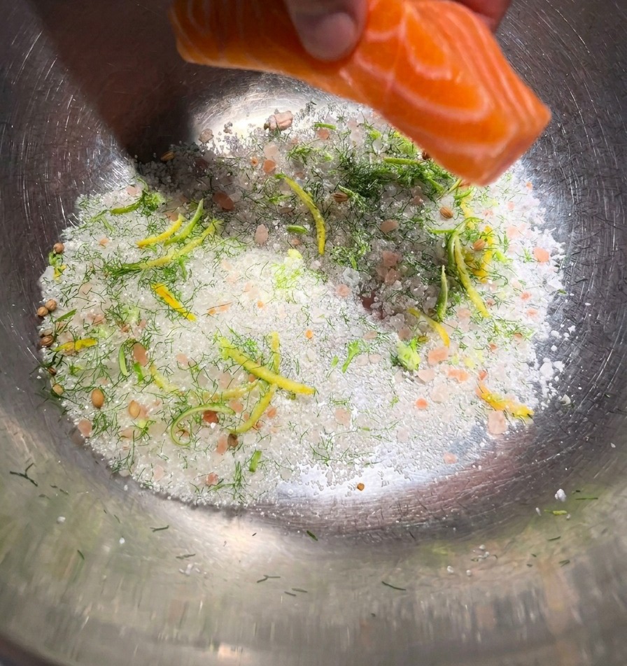 Une main déposant un filet de saumon orange vif sur un lit de sel, de sucre et dherbes dans un bol de préparation.