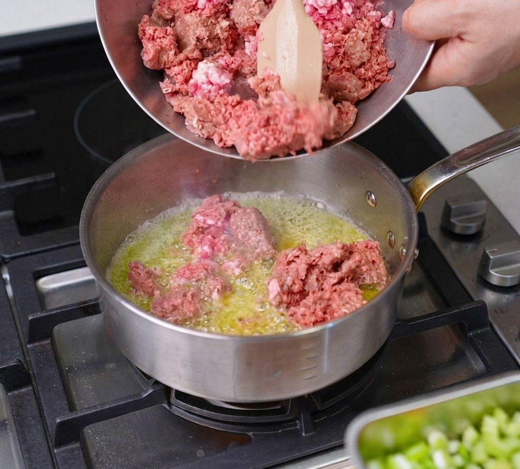 Du bœuf et du porc hachés versés depuis un bol métallique dans une poêle chaude contenant du beurre fondu.