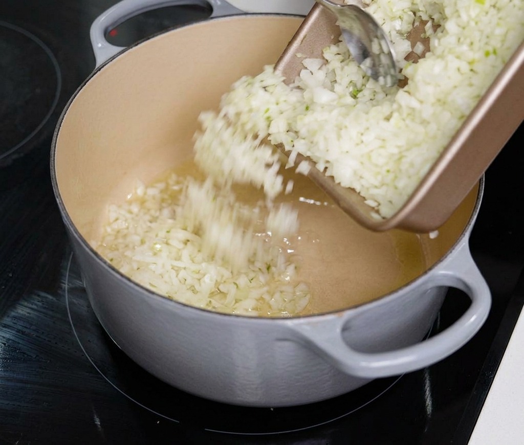Des oignons blancs finement émincés versés depuis un plateau rectangulaire dans une grande cocotte grise.