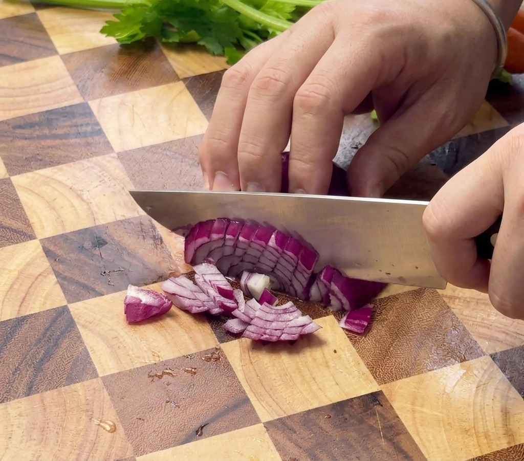 Gros plan sur des mains utilisant un couteau de chef pour couper un oignon rouge sur une planche en bois.