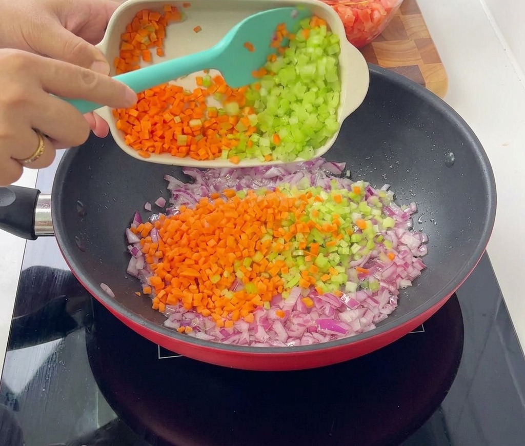 Une personne versant un mélange de carottes oranges et de céleri vert en dés dans une grande poêle rouge contenant les oignons violets sautés.