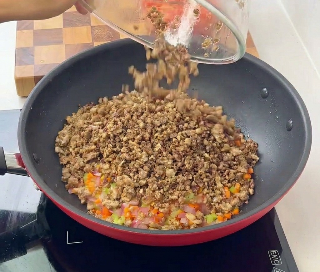 Viande hachée cuite versée dun bol en verre dans une poêle contenant un mélange de légumes sautés en dés.