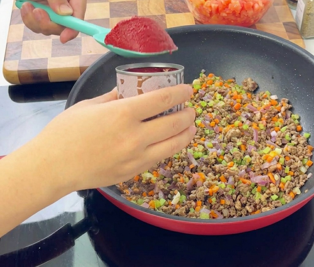 Une spatule turquoise tenant une cuillerée de concentré de tomate rouge foncé au-dessus dune poêle remplie de viande dorée et de légumes en dés.
