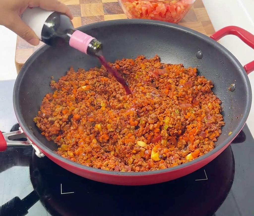 Du vin rouge versé dune bouteille dans une poêle rouge contenant les ingrédients de la sauce à la viande.