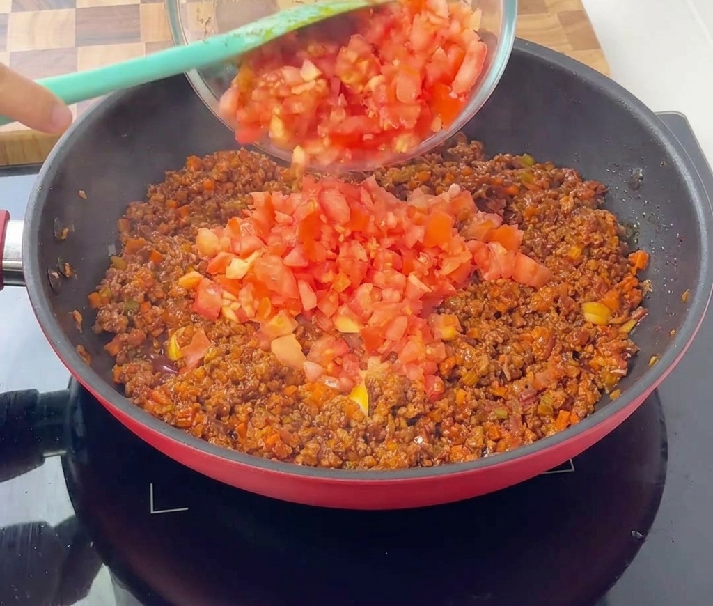 Des tomates concassées versées dun bol en verre transparent dans une poêle de sauce à la viande et aux légumes qui mijote.