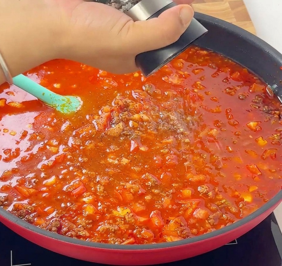 Une main tenant un moulin à poivre et ajoutant du poivre noir frais dans une poêle de sauce à la viande et à la tomate qui mijote.