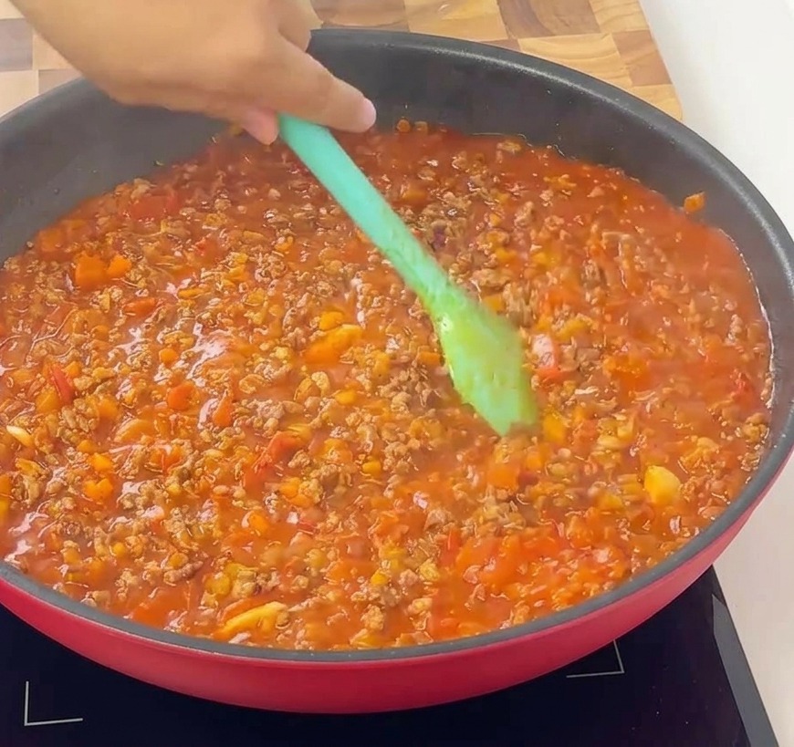 Une spatule vert clair mélangeant une sauce bolognaise épaisse, riche et parfaitement cuite dans une poêle rouge.