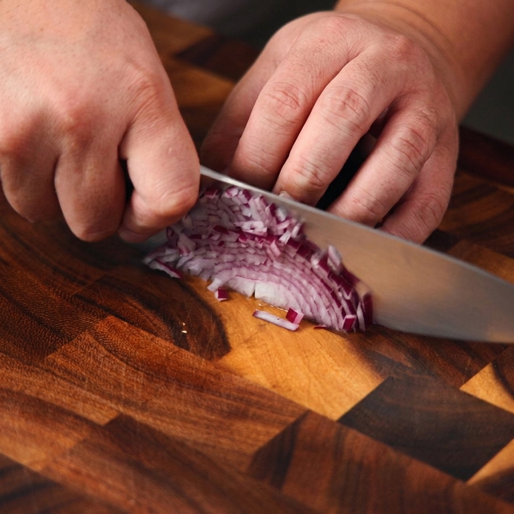Gros plan sur des mains utilisant un couteau de chef pour émincer finement un oignon rouge sur une planche en bois.