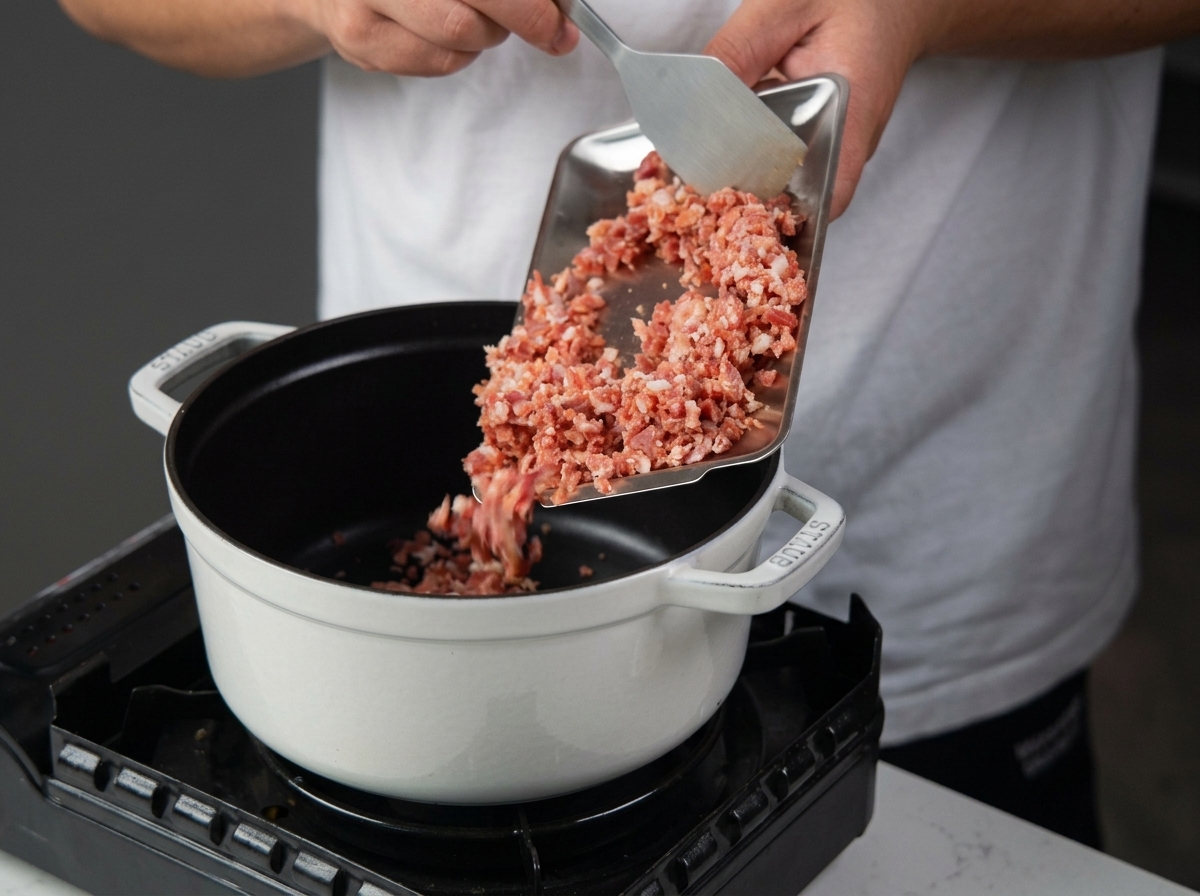 Versement de dés de pancetta depuis un plateau métallique dans une cocotte en fonte émaillée blanche posée sur un réchaud.
