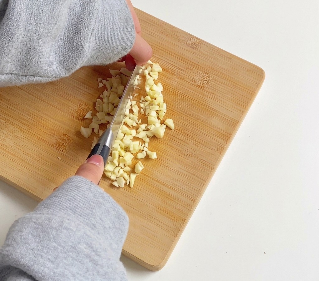 Une personne utilisant un couteau de cuisine argenté pour émincer finement plusieurs gousses dail sur une planche à découper en bambou.