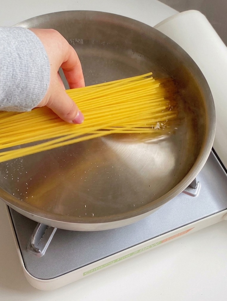 Une main plongeant un nid de spaghetti secs dans une casserole en acier inoxydable remplie deau frémissante, de gouttes dhuile et de sel.