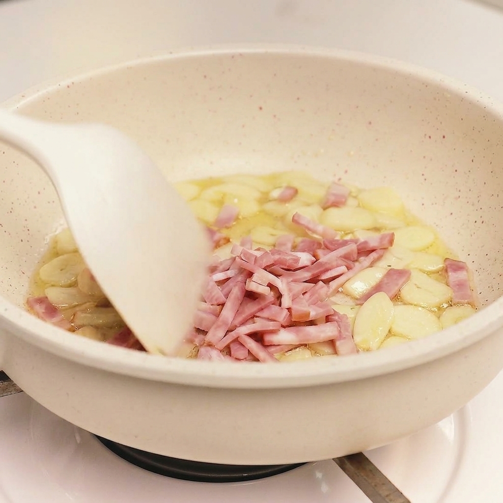 Une spatule blanche mélangeant le bacon en morceaux et lail dans une poêle de couleur claire.