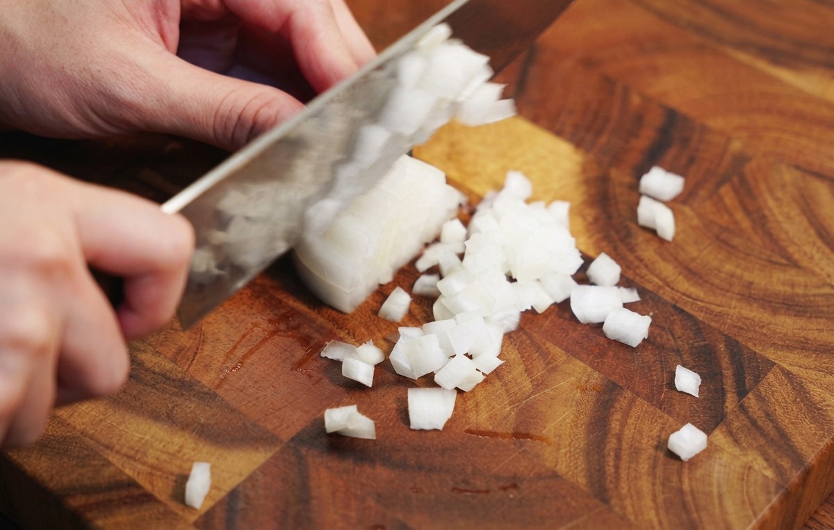Mains utilisant un couteau de chef pour émincer un oignon blanc sur une planche à découper en bois.