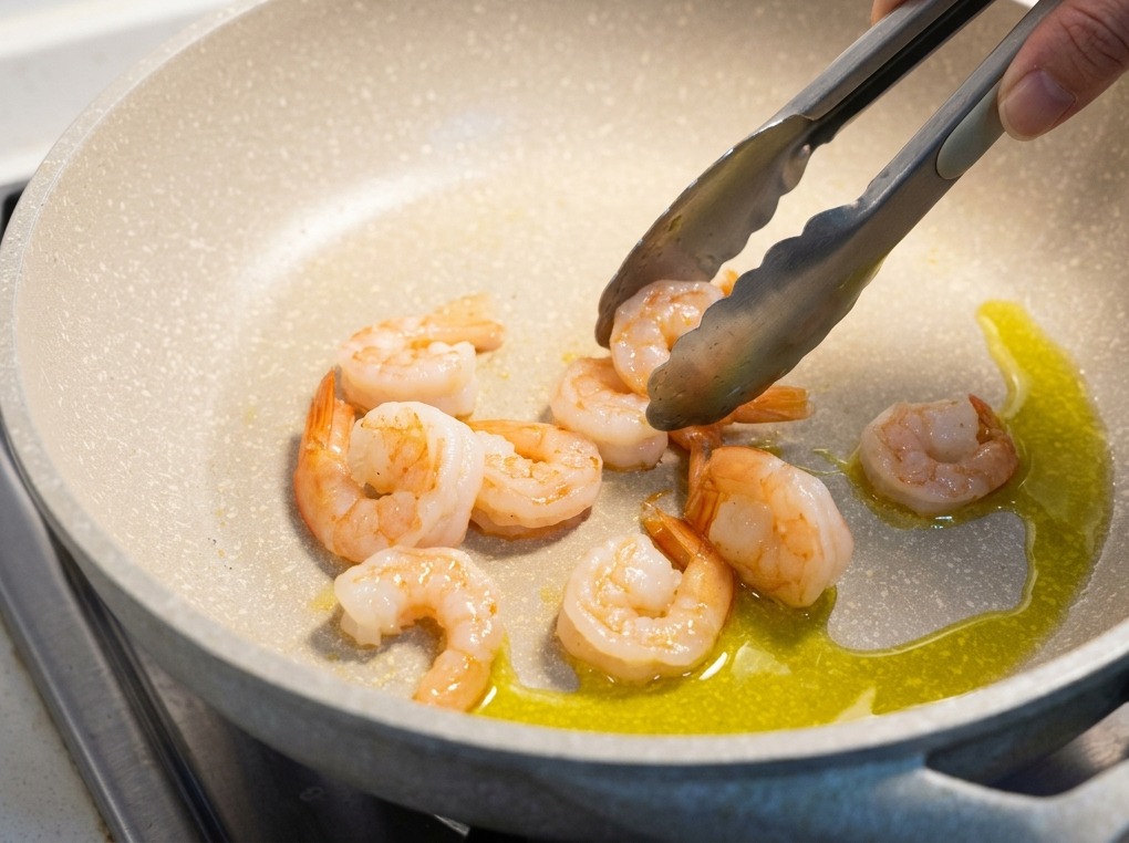 Crevettes roses cuites retournées avec une pince métallique dans une poêle contenant de lhuile dolive.