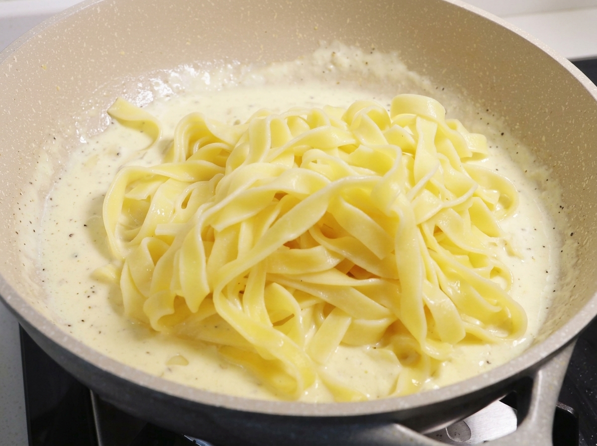 Un dôme de fettuccine cuites déposé sur une sauce à la crème blanche frémissante dans une grande poêle.