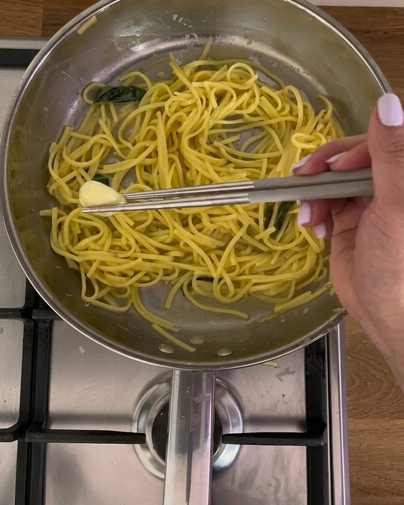 Des baguettes de cuisine en métal retirant une gousse dail fondante dune poêle en acier remplie de pâtes jaunes et de basilic.