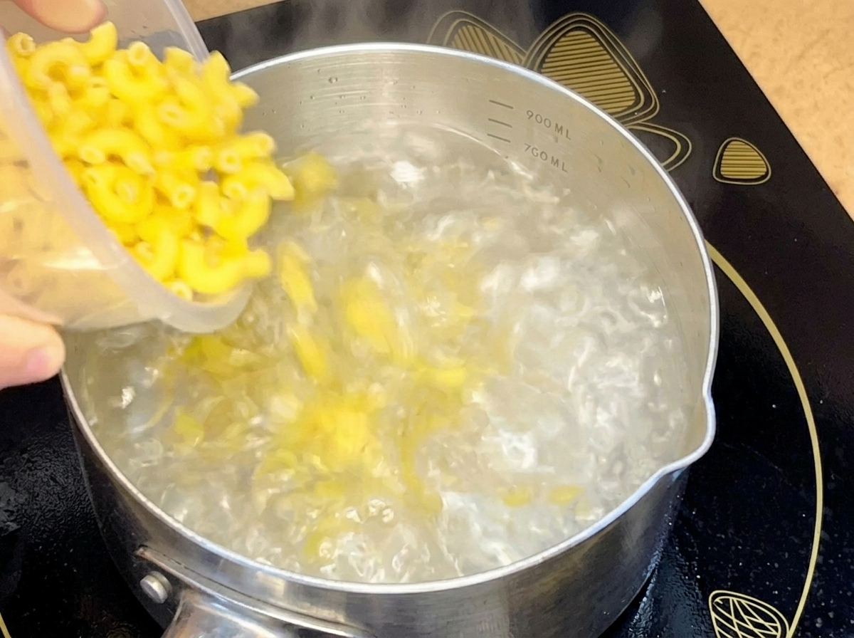 Macaronis crus versés depuis un récipient dans une casserole en acier inoxydable remplie deau bouillante.