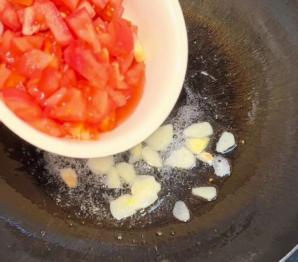 Dés de tomates versés depuis un bol blanc dans une poêle contenant de lhuile chaude et des lamelles dail dorées.