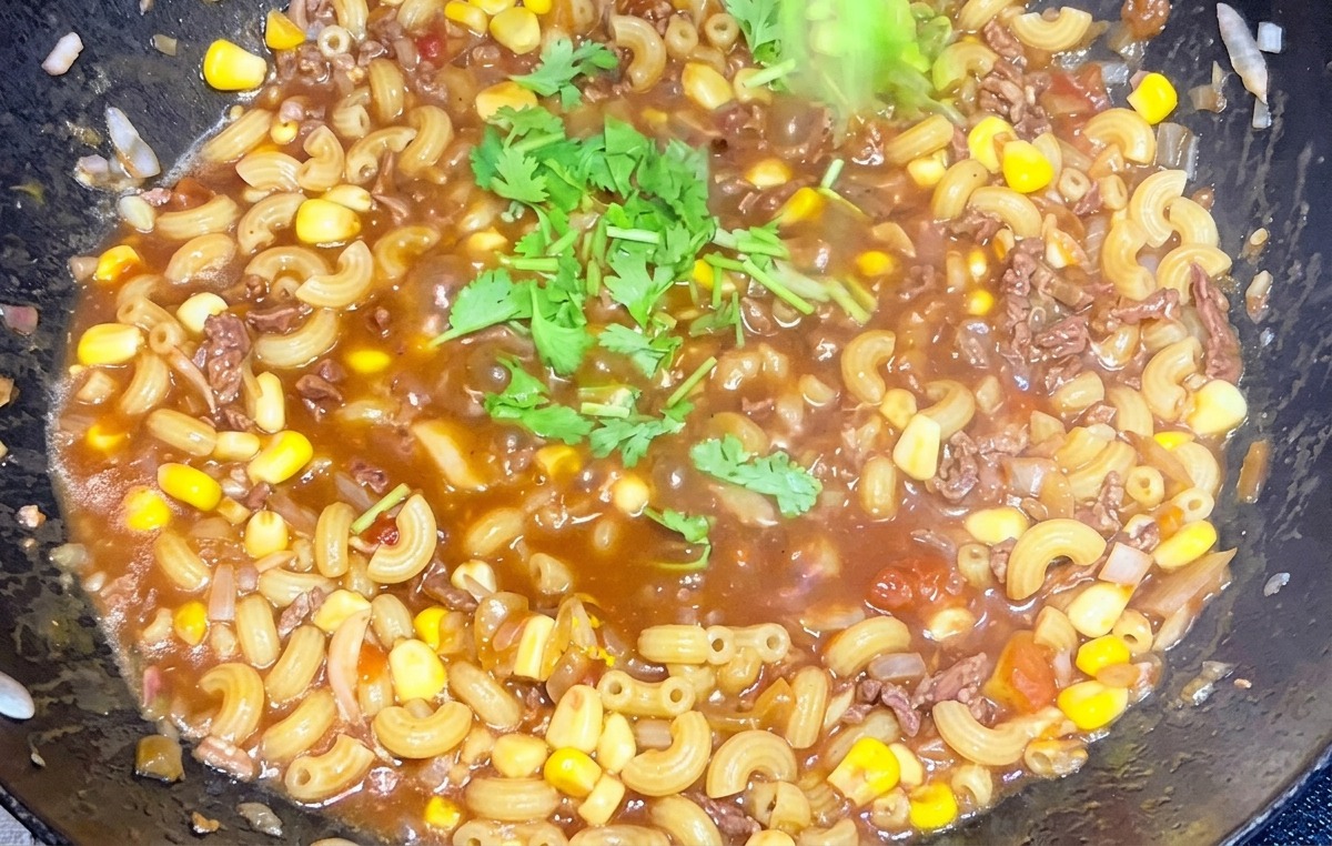 Une main parsemant de la coriandre verte hachée sur une poêle fumante de macaronis au bœuf et à la tomate.