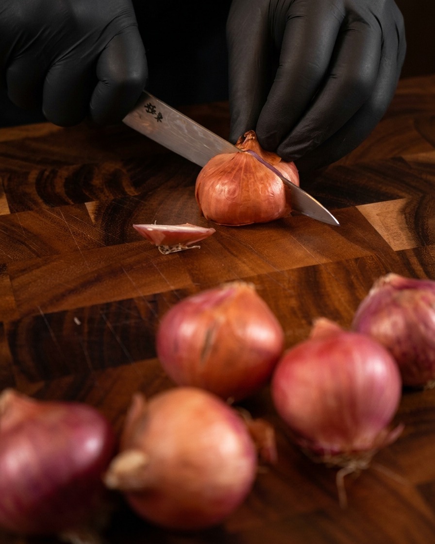 Mains portant des gants noirs utilisant un couteau de chef bien aiguisé pour émincer finement des échalotes entières sur une planche en bois sombre.