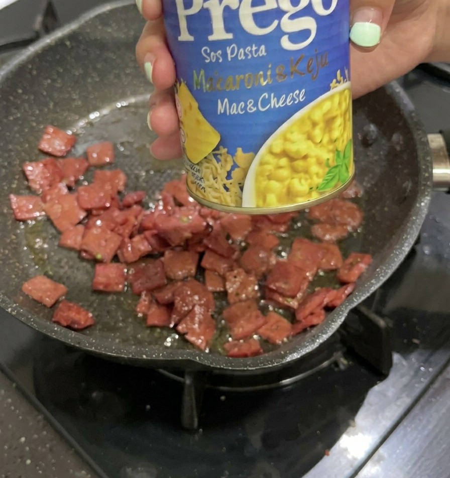 Une main tenant une conserve bleue et jaune de sauce pour pâtes au fromage Prego au-dessus dune poêle contenant des morceaux de salami grillés.