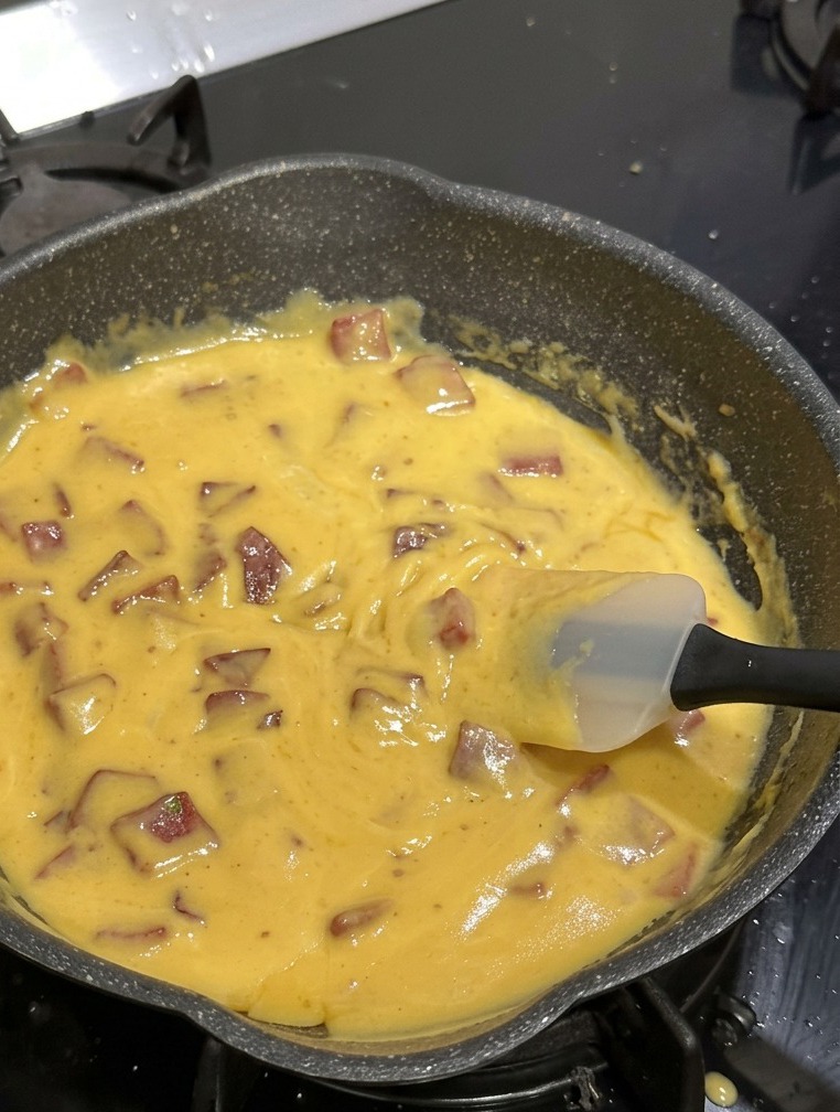 Une poêle antiadhésive noire remplie de macaronis, de dés de salami de bœuf et dune sauce au fromage jaune vif et épaisse, remuée avec une spatule en silicone blanche.