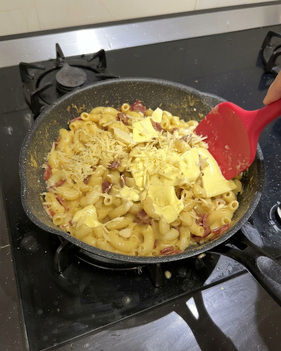 Des macaronis, du salami, des tranches de fromage et du fromage râpé dans une poêle noire, remués avec une spatule en silicone rouge.