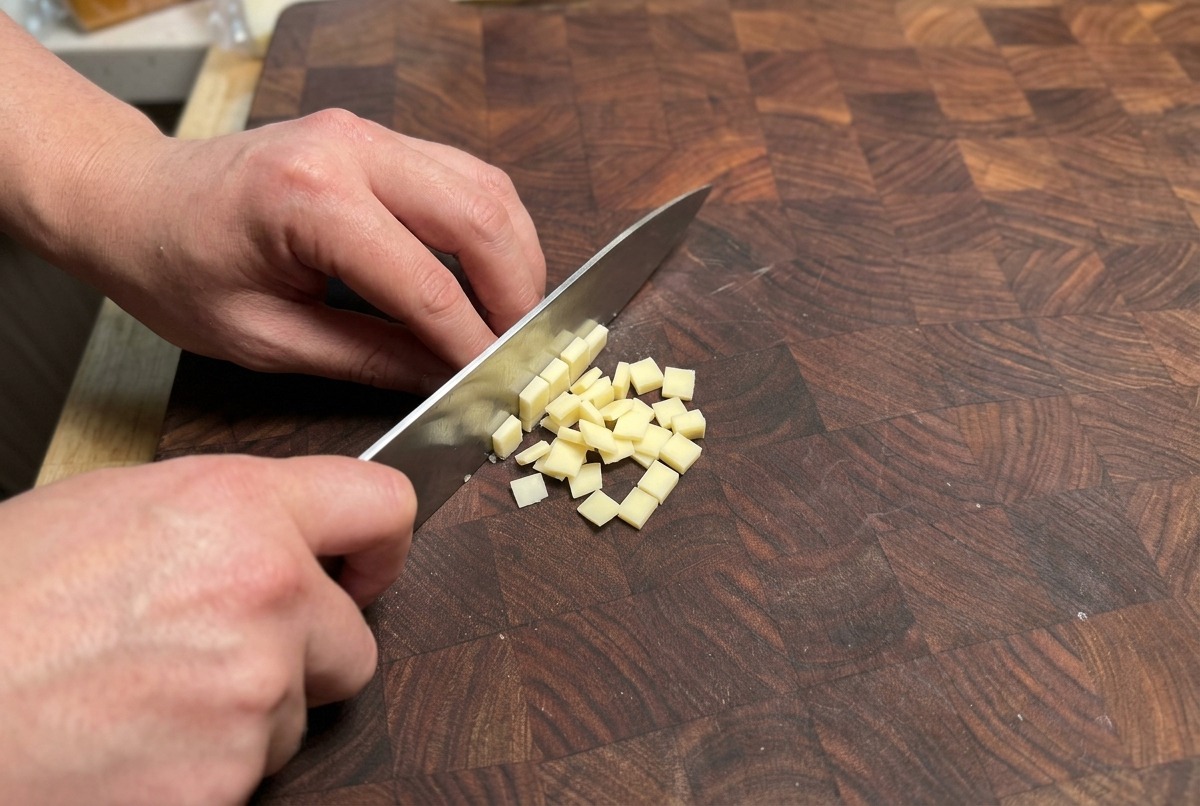 Mains utilisant un couteau de chef pour couper un bloc de fromage jaune pâle en petits cubes sur une planche à découper en bois.