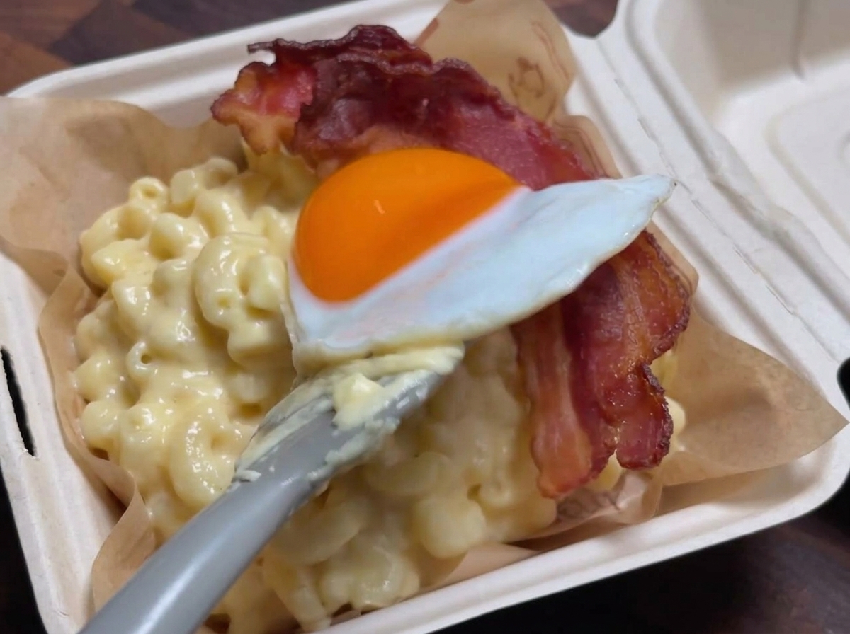 Une spatule déposant délicatement un œuf au plat sur une bande de bacon croustillant posée sur une portion de macaronis au fromage crémeux.